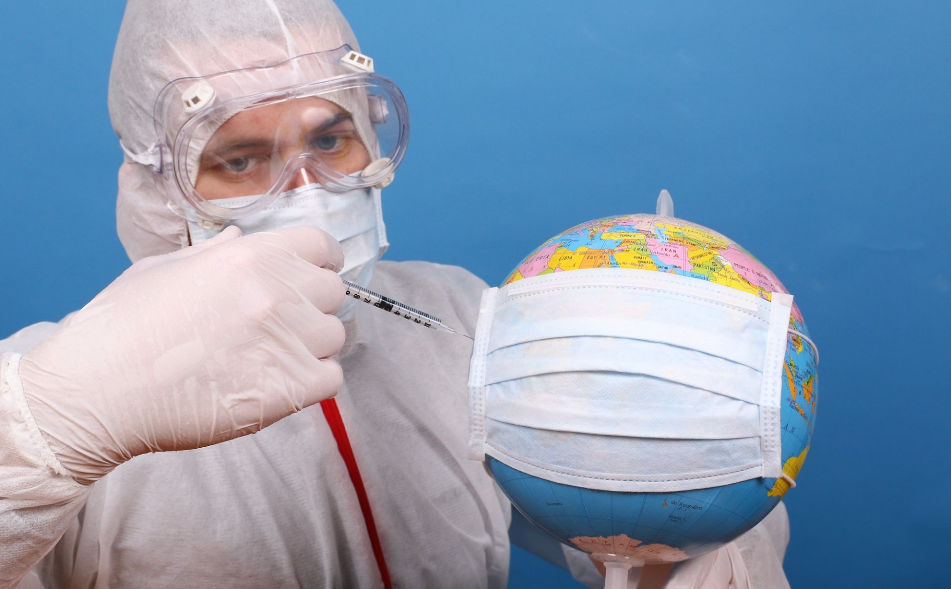 A person dressed in a hazmat suit injects a syringe into a globe covered that is covered with a face mask