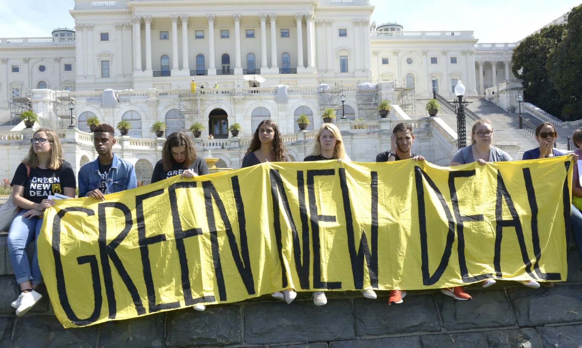 Eight youth hold a large yellow banner that read Gree New Deal in front of the Capitol Building